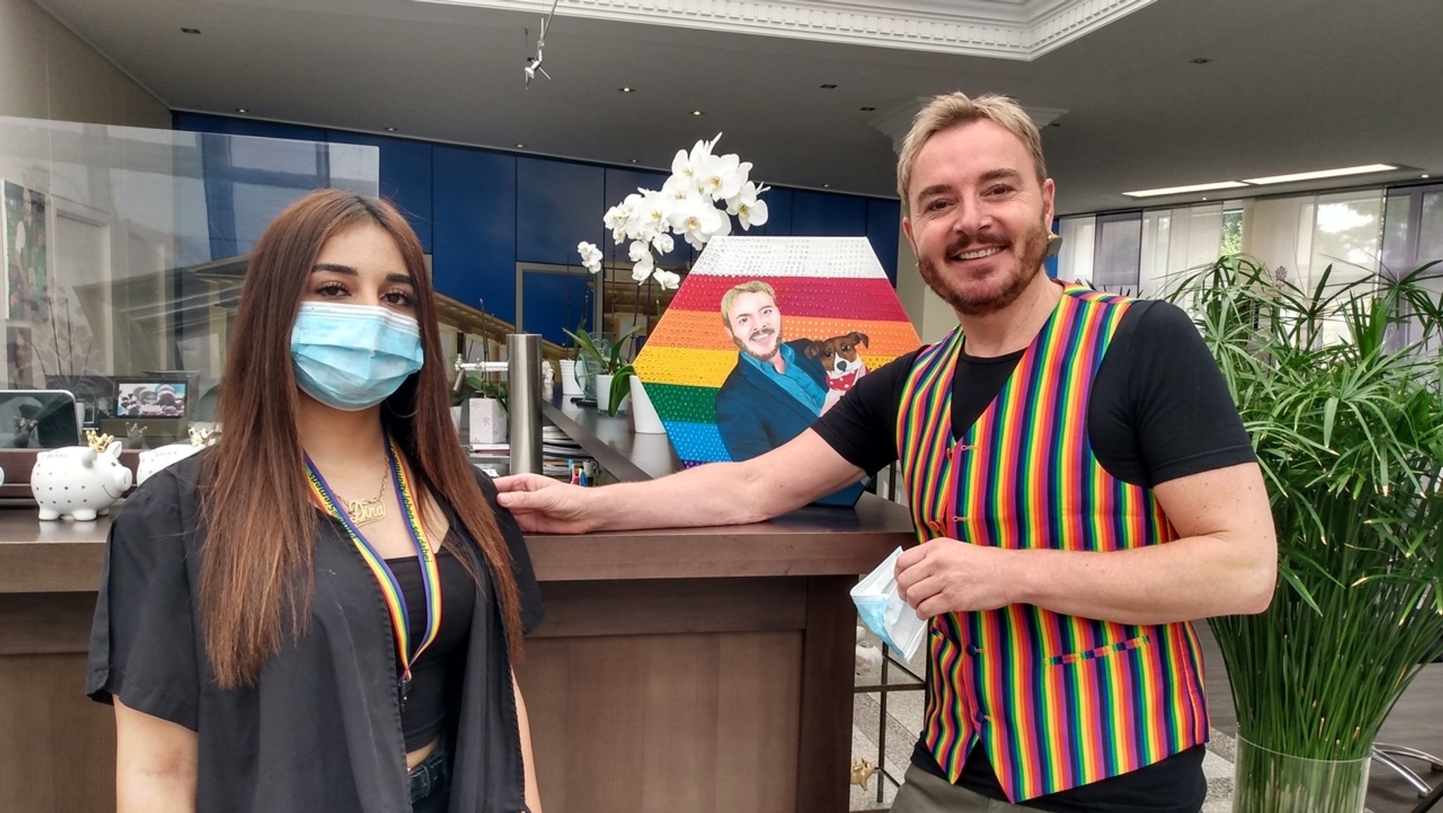 Ein Mann in Regenbogenweste und eine Frau mit Regenbogen-Schlüsselband in einem Friseursalon.