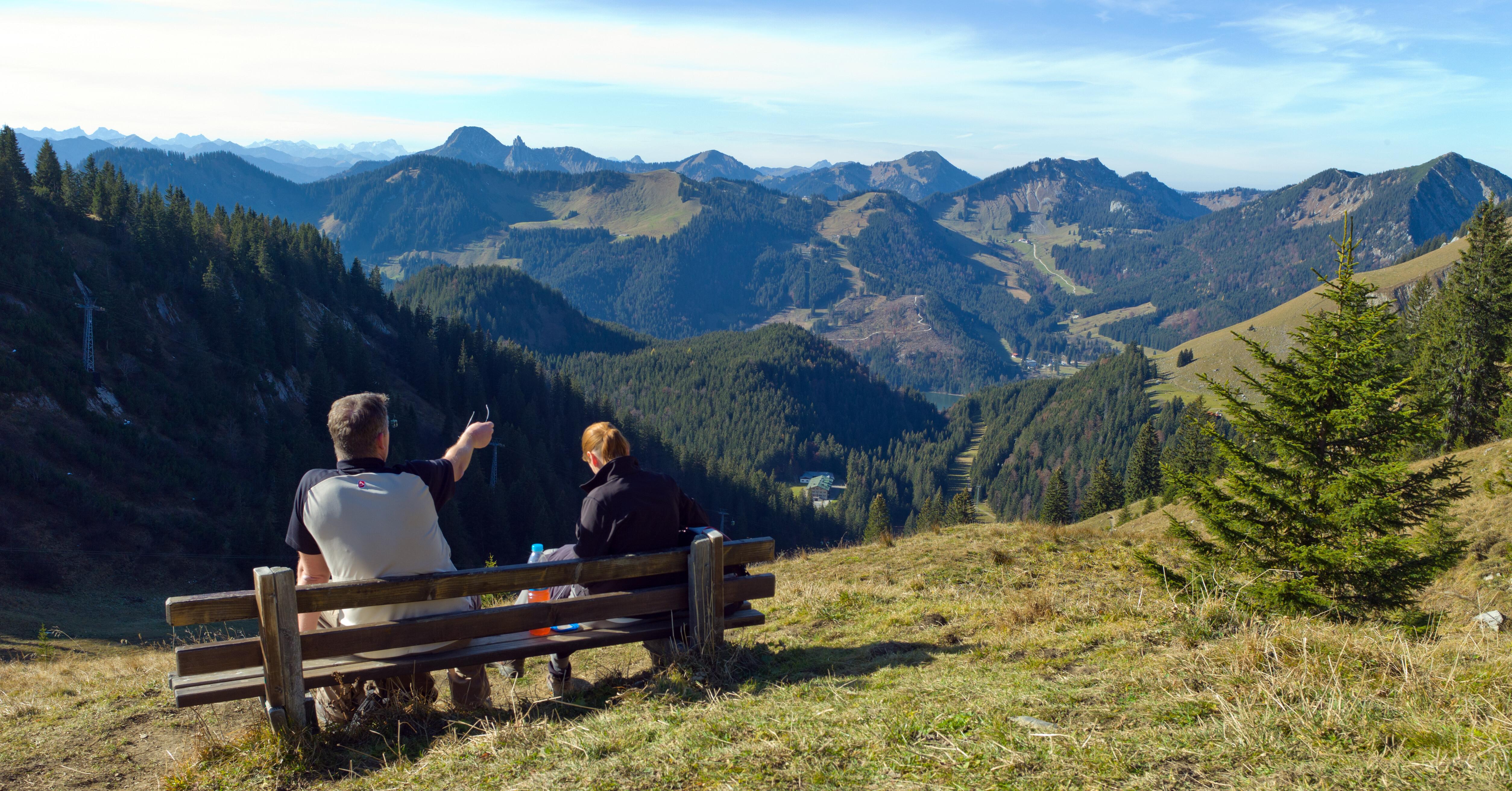 Zwei Wanderer sitzen oberhalb des Spitzingsees mit Aussicht auf das Alpenpanorama auf einer Bank. 