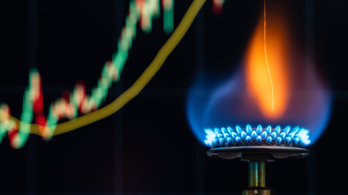 Eine bläuliche Flamme aus einem Gaskocher ist in einem Büro vor einem Bildschirm eines Computers zu sehen, auf dem der Preis von Erdgas als Kerzenchart dargestellt wird.