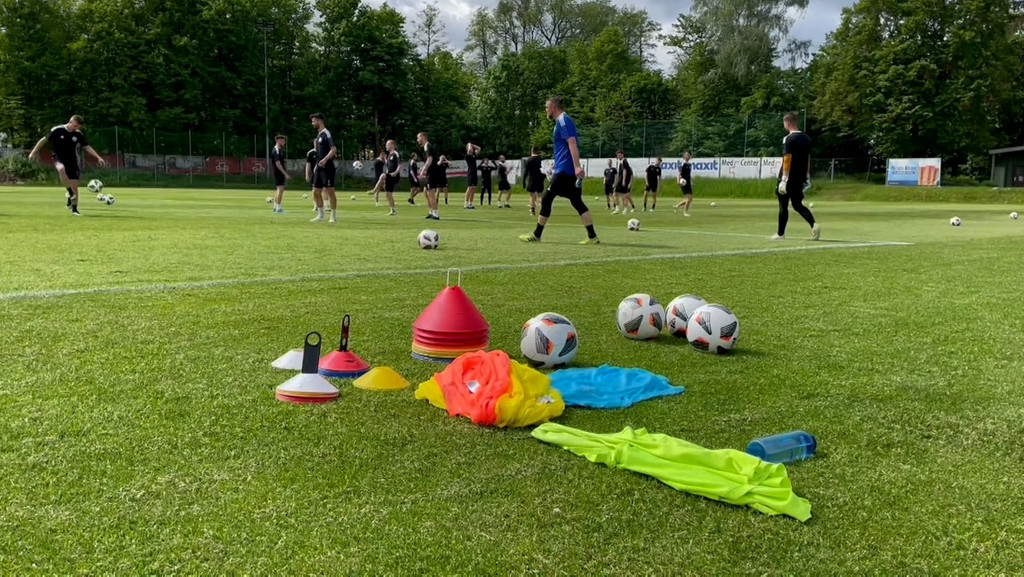 Auf dem Fußballplatz trainieren mehrere Spieler, am Spielfeldrand liegen unter anderem mehrere Bälle.