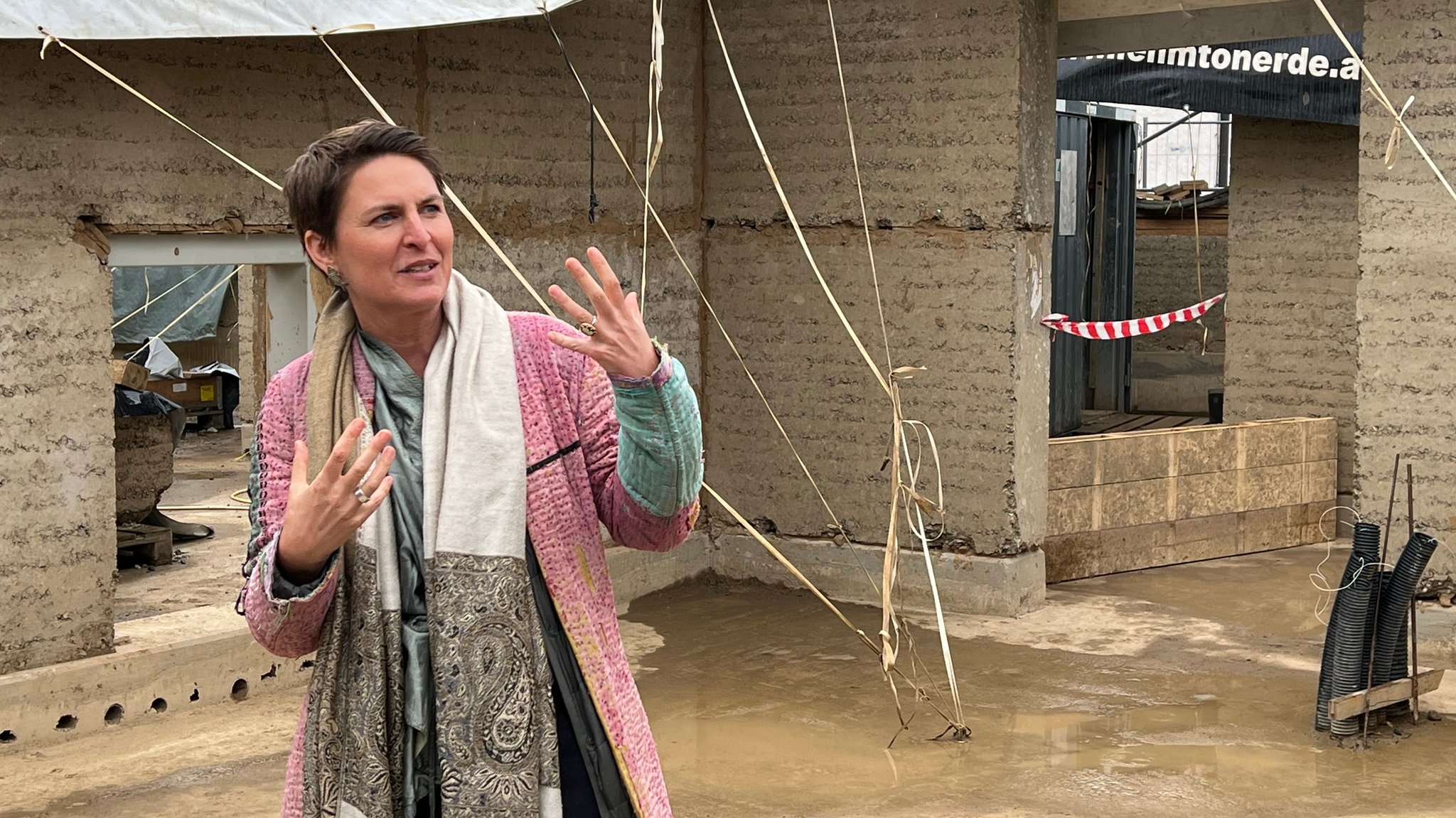 Architektin Anna Heringer gestikuliert auf einer Baustelle. Im Hintergrund sieht man dicke Mauern aus geschichtetem und verdichtetem Lehm. 