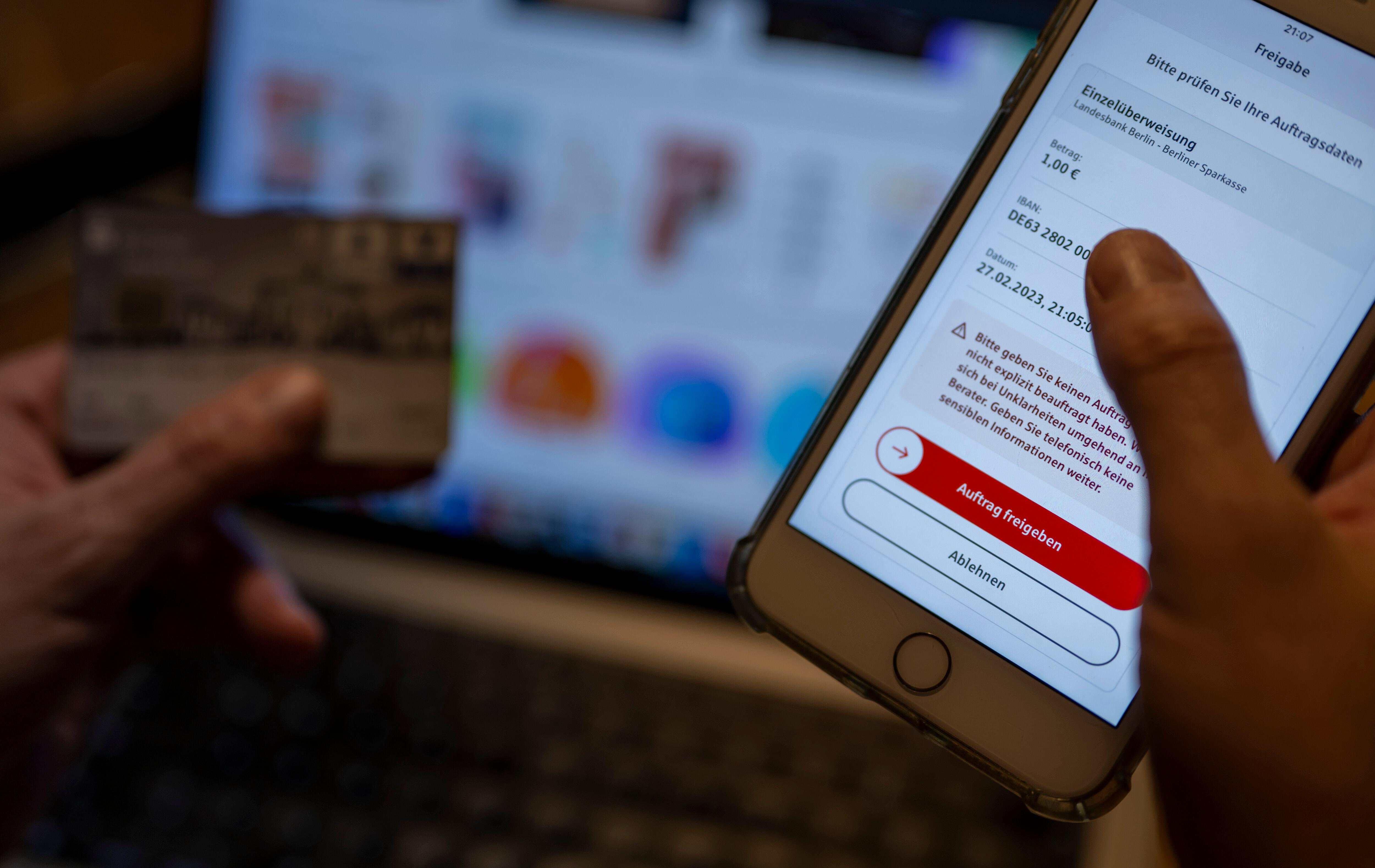 (Symbolbild) Ein Mann überweist in einer Banking-App auf einem Smartphone Geld, während im Hintergrund auf einem Laptop die Seite eines Online-Handels angezeigt wird.