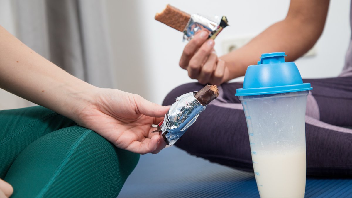 Zu sehen sind zwei sitzende Frauen, von denen man nur Beine in Sportbekleidung und die Hände sieht. jede hält einen Proteinriegel. Davor ein Behältnis mit einem Proteindrink (gestellte Szene).