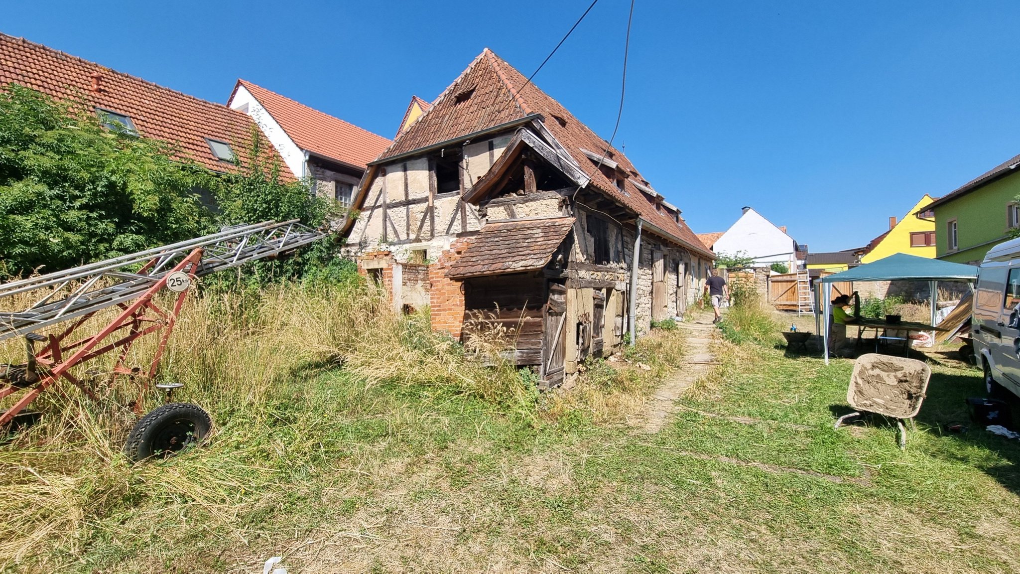 Historisches Fachwerkhaus mit starkem Sanierungsbedarf. Freigelegtes Fachwerk, beschädigte Fassade und überwuchertes Grundstück. Im Vordergrund Baustellengeräte, rechts ein Pavillon und zwei Personen bei Arbeiten. Sonniger Tag, Dorfbebauung im Hintergrund.