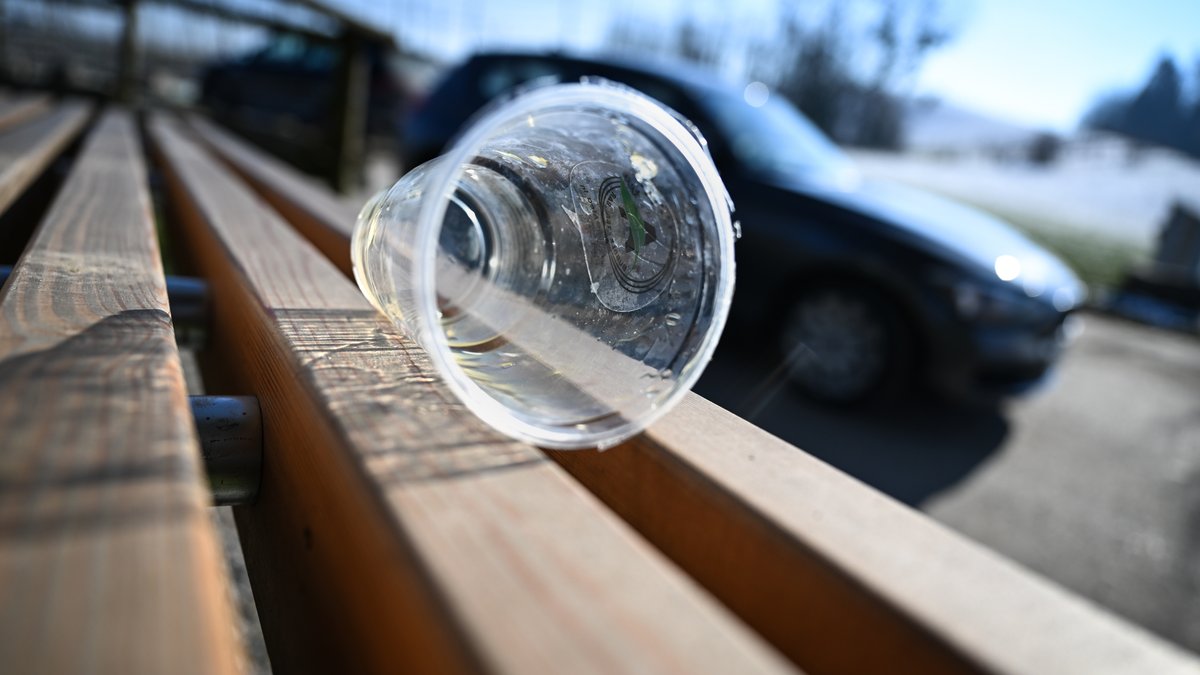 Ein transparenter Plastikbecher liegt auf einer Rastbank, während ein Auto vorbei fährt.