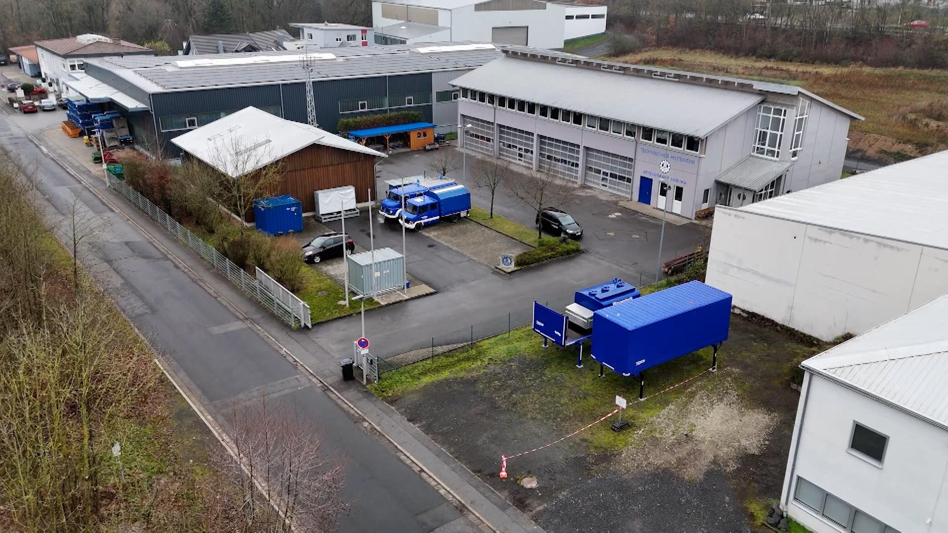 Vor Zweckbauten parken blaue Nutzfahrzeuge, betrachtet aus der Vogelperspektive.