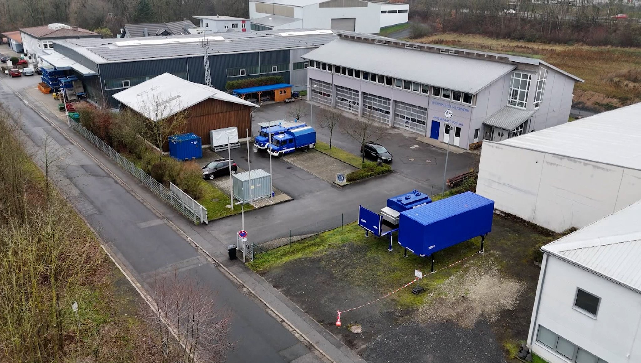 Vor Zweckbauten parken blaue Nutzfahrzeuge, betrachtet aus der Vogelperspektive.