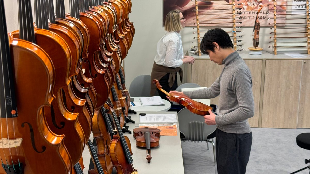 Geigen und Chellos stehen aufgereiht auf einem Tisch, ein Mann hält eine Geige in der Hand.