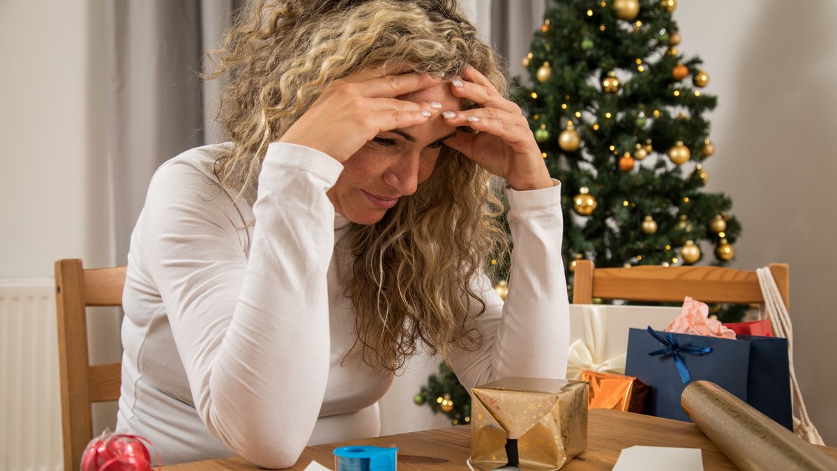 Eine Frau schaut gestresst auf Geschenke und Verpackungen. Im Hintergrund steht ein Weihnachtsbaum.