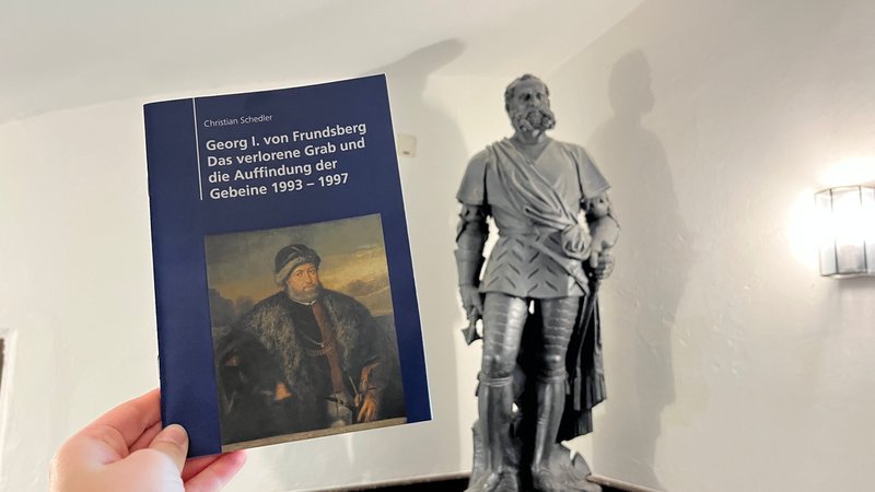 Im Vordergrund sieht man hochgehalten die Broschüre "Georg I. von Frundsberg: Das verlorene Grab und die Auffindung der Gebeine 1993 bis 1997" inklusive Porträt von Frundsberg. Im Hintergrund ist eine Statue von ihm sichtbar.