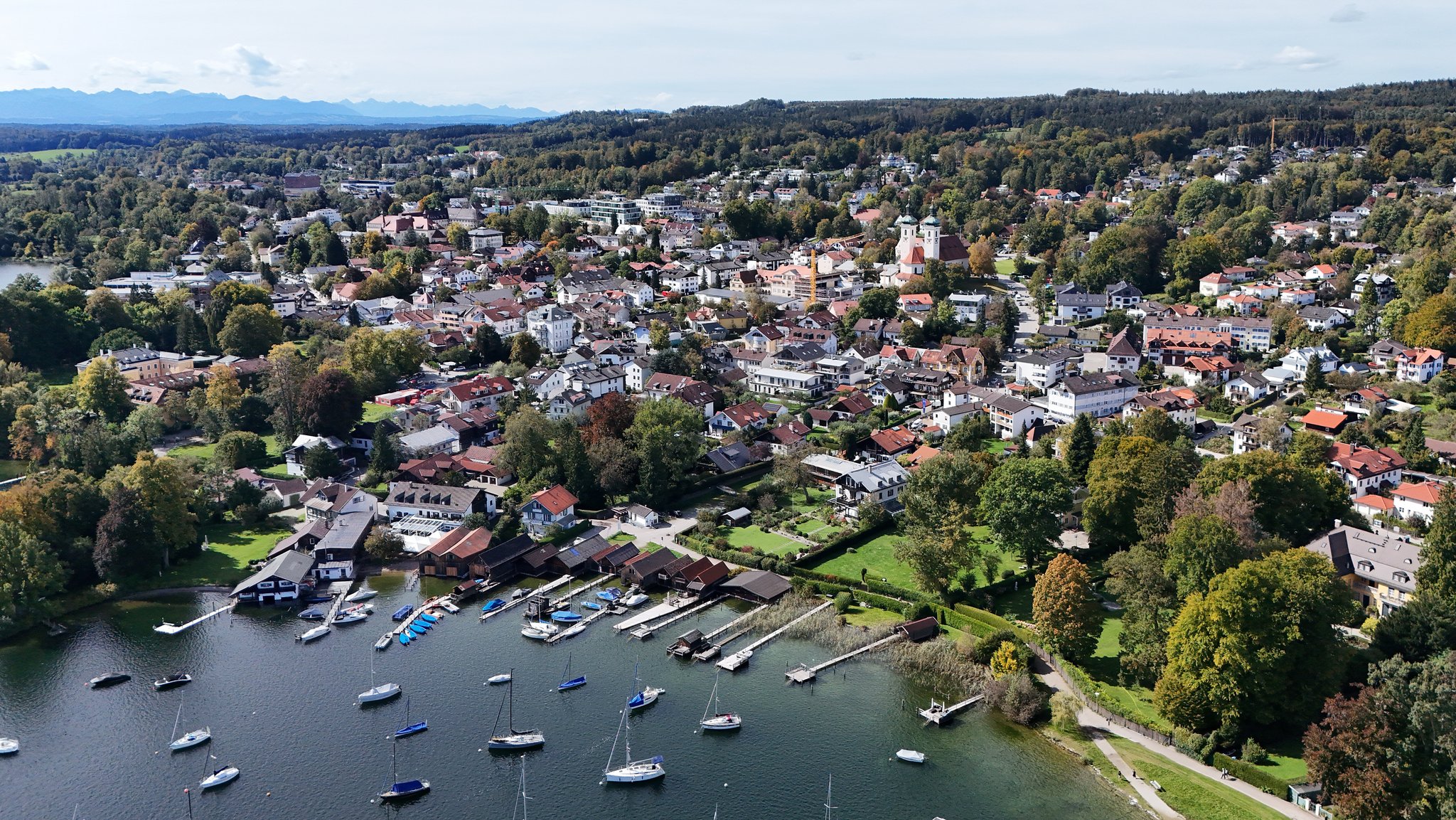 Blick aus der Vogelperspektive auf die Brahmspromenade in Tutzing am Starnberger See. Am unteren Bildrand liegen Segelboote im Wasser, am oberen Bildrand sieht man die Alpenkette, dazwischen breitet sich der Ort Tutzing aus. (Archivbild)