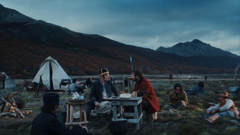 Eine Handvoll Männer sitzt in weiter, leerer Landschaft verstreut um einen Tisch, daneben lodert ein Lagerfeuer. 