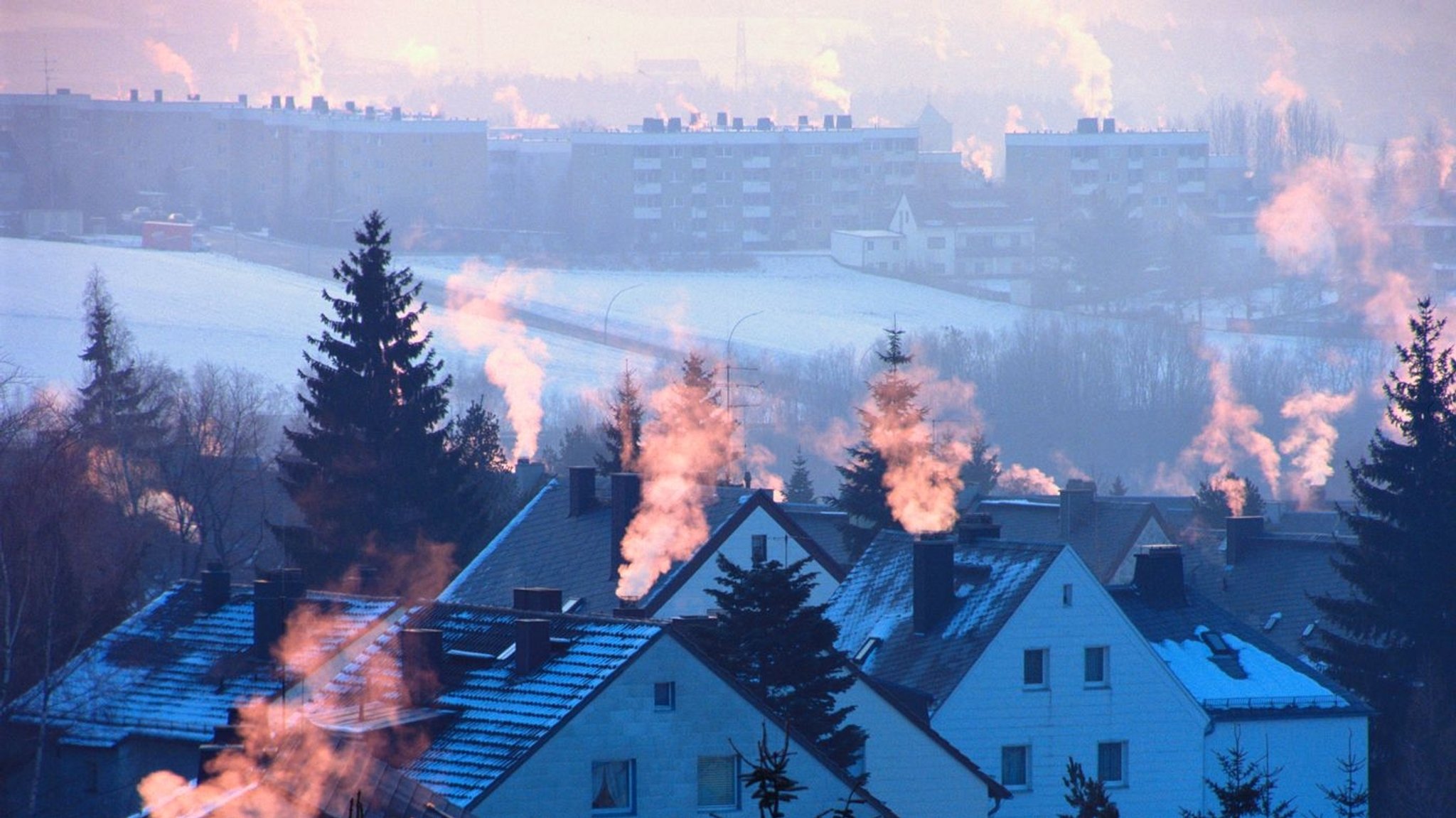 Abgase aus Schornsteinen auf schneebedeckten Dächern werden von der niedrigstehenden Sonne angestrahlt.