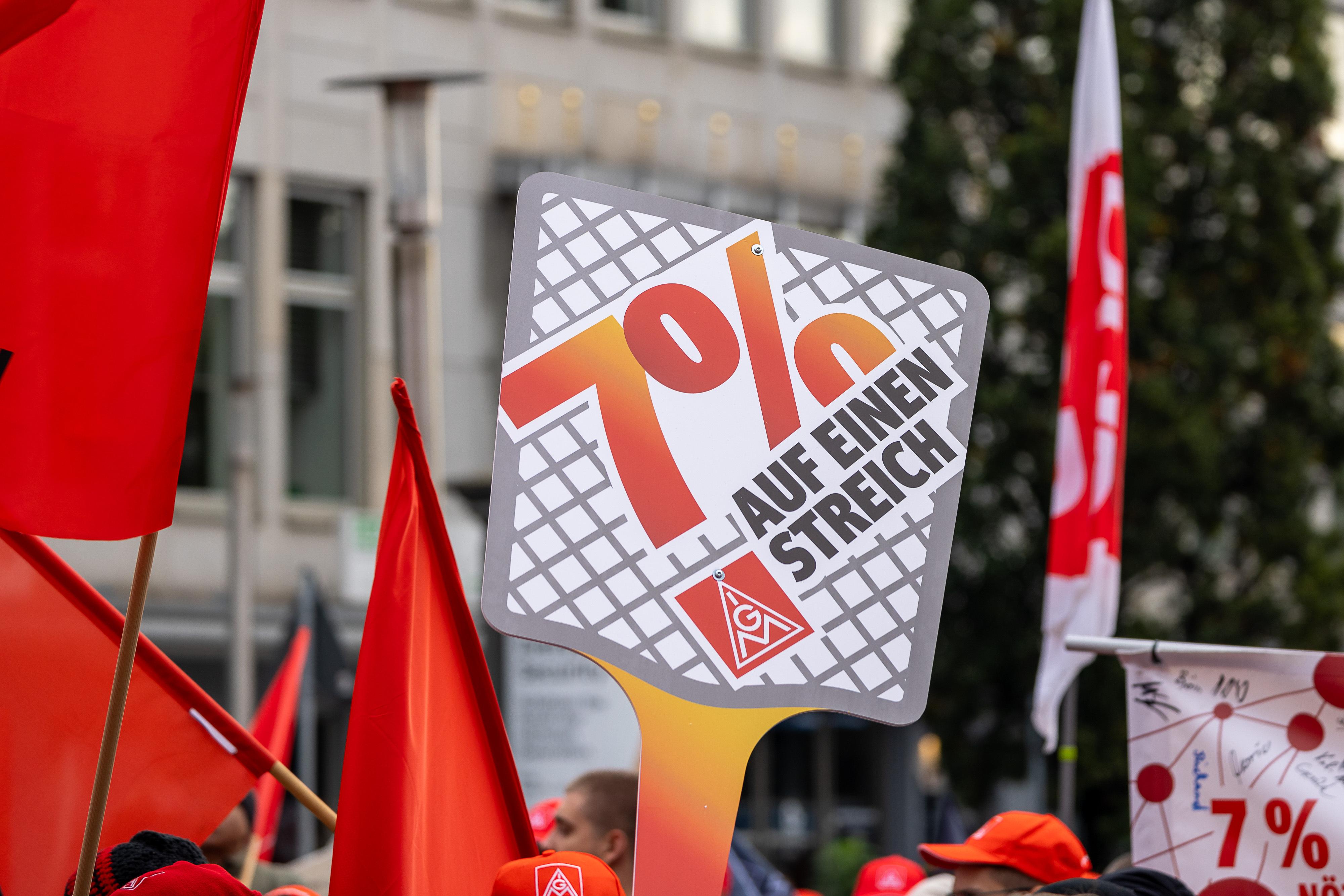 Ein Schild mit der Aufschrift "Sieben Prozent" wird hochgehalten, rote Fahnen und Mützen im Hintergrund