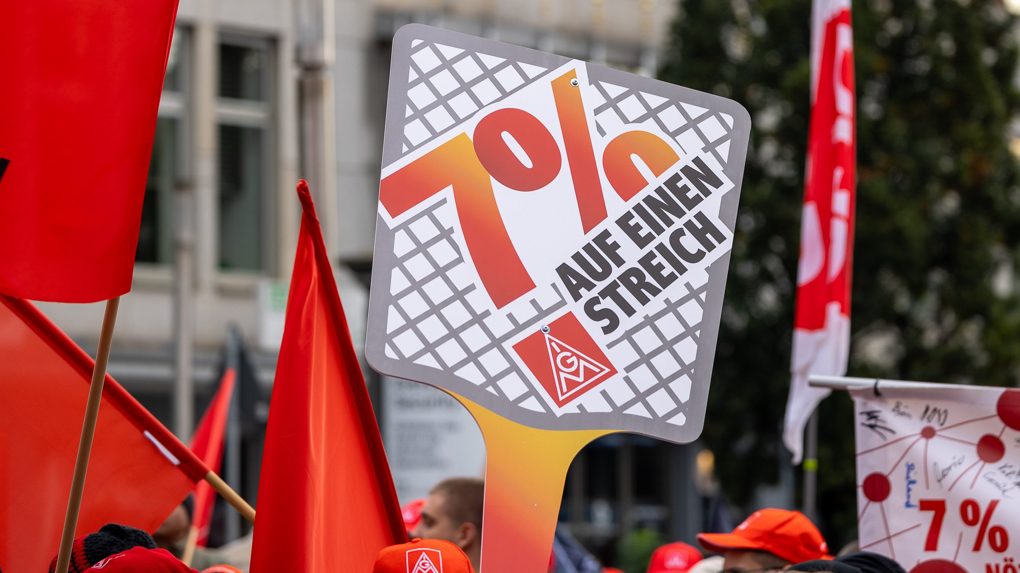 Ein Schild mit der Aufschrift "Sieben Prozent" wird hochgehalten, rote Fahnen und Mützen im Hintergrund