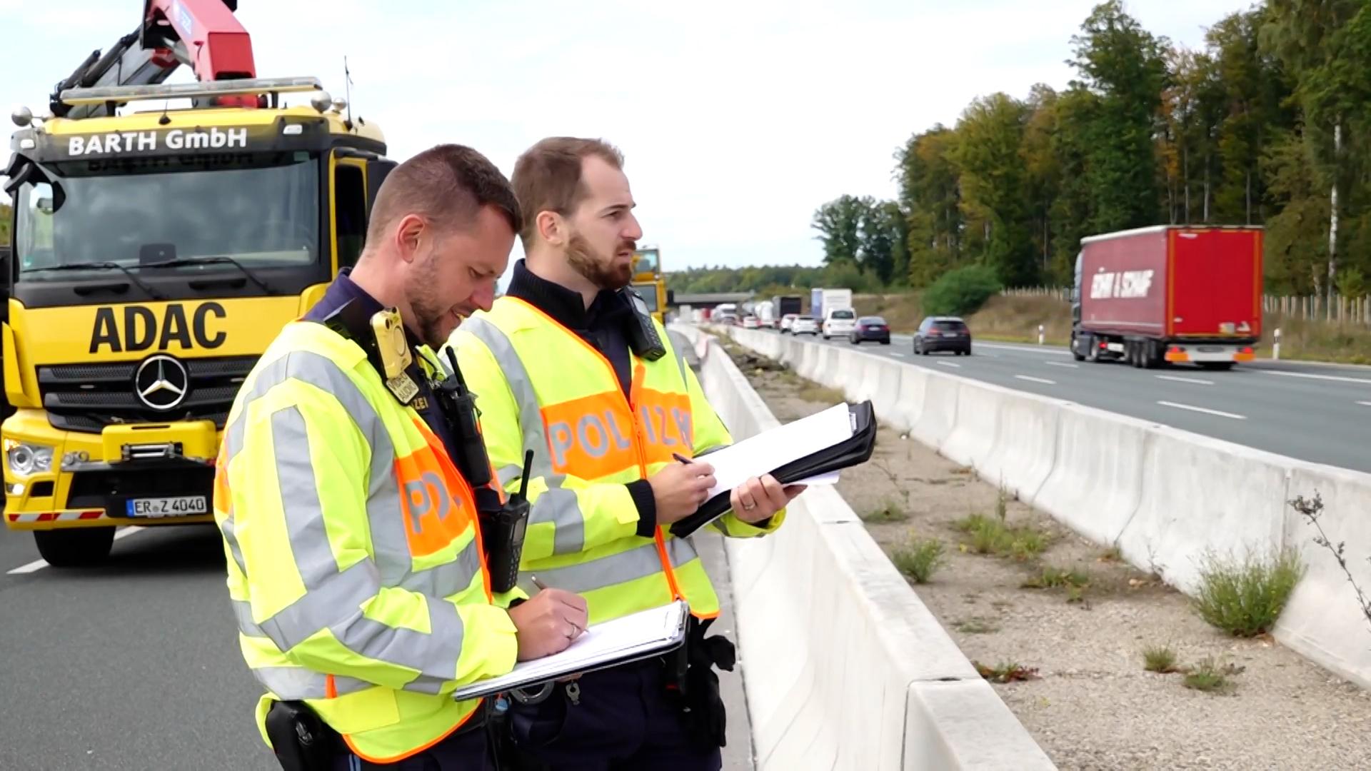 Zwei Polizisten in Warnwesten schauen mit Klemmbrettern und Papieren in der Hand auf die Gegenfahrbahn der A3.
