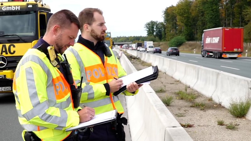 Zwei Polizisten in Warnwesten schauen mit Klemmbrettern und Papieren in der Hand auf die Gegenfahrbahn der A3. | Bild: News5 Zwei Polizisten in Warnwesten schauen mit Klemmbrettern und Papieren in der Hand auf die Gegenfahrbahn der A3.