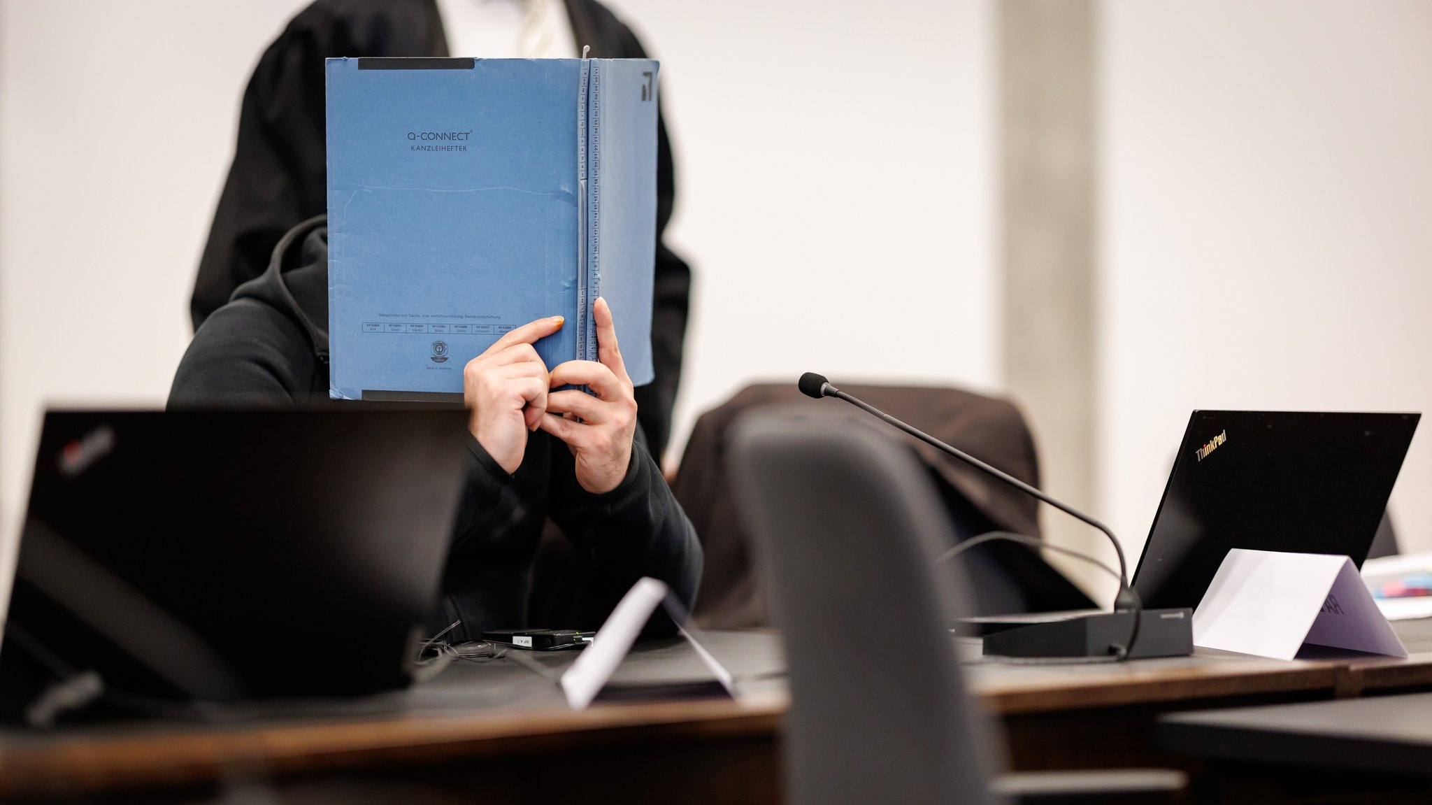 Ein Mann sitzt mit einem blauen Aktenbinder vor dem Gesicht im Gerichtssaal.