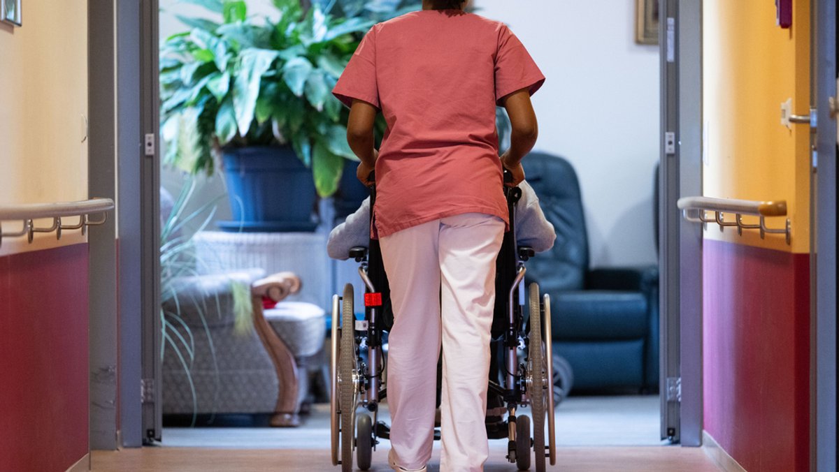 Eine Bewohnerin eines Pflegeheims wird im Rollstuhl von einer Pflegerin einen Gang entlang geschoben.