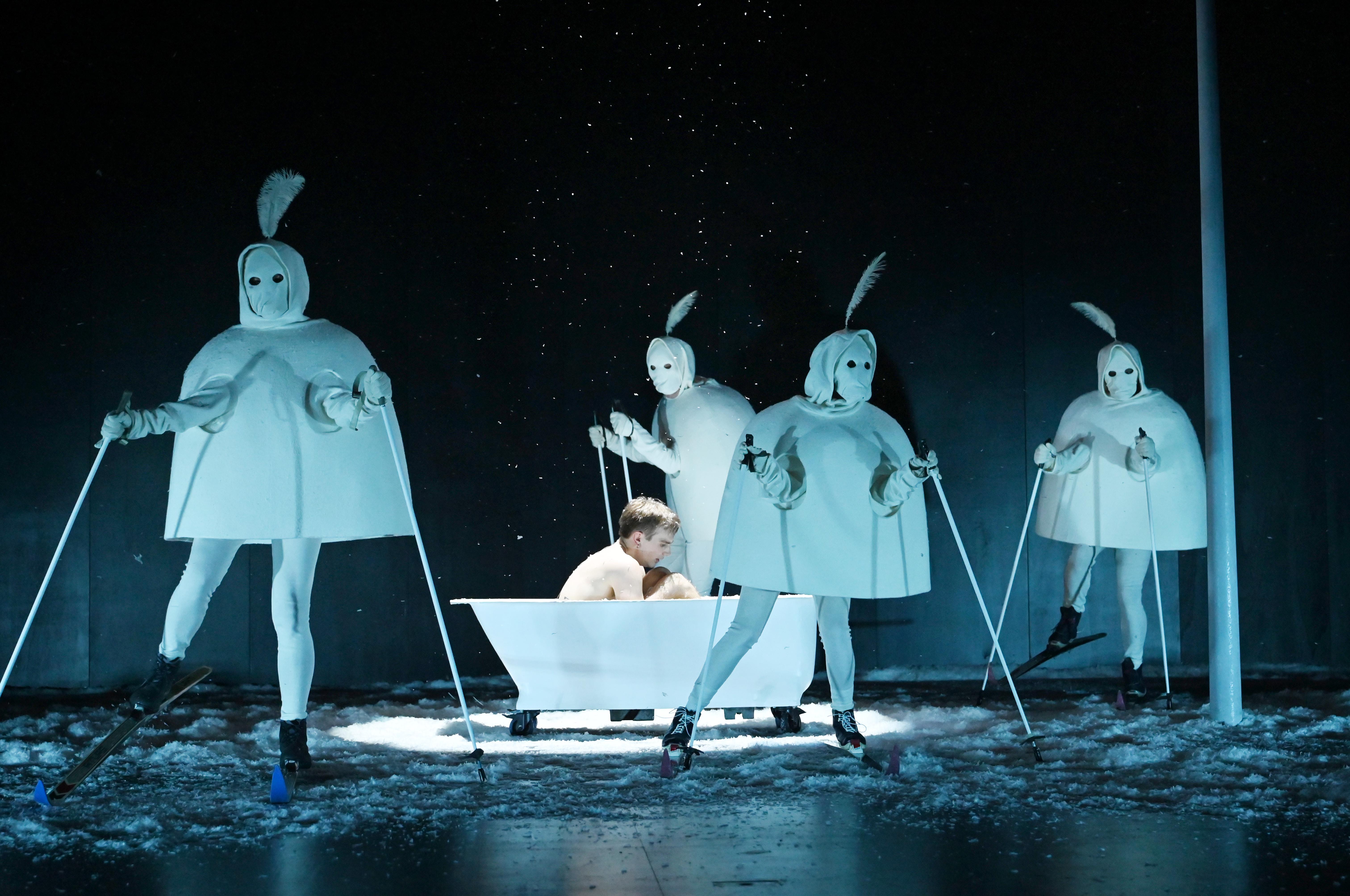 Zu sehen: Figur Behrendt in einer Badewanne, um ihn herum seltsame Gestalten in weißem Kostüm mit Skier auf Kunstschnee.