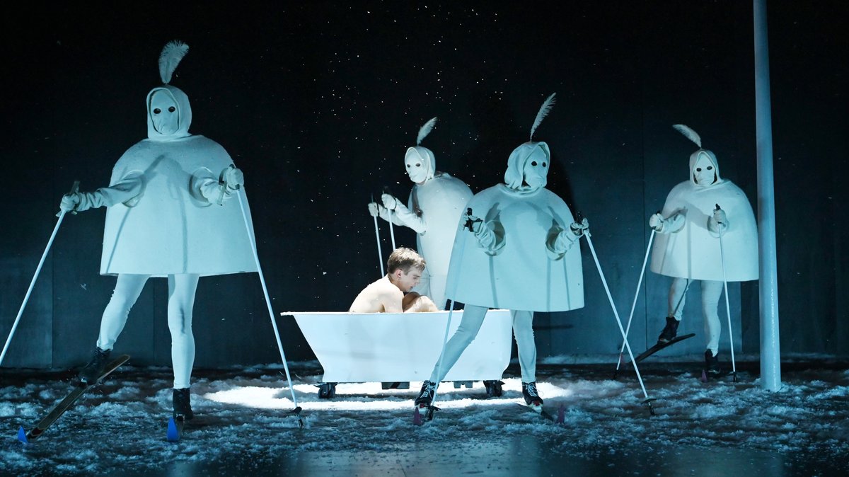 Zu sehen: Figur Behrendt in einer Badewanne, um ihn herum seltsame Gestalten in weißem Kostüm mit Skier auf Kunstschnee.