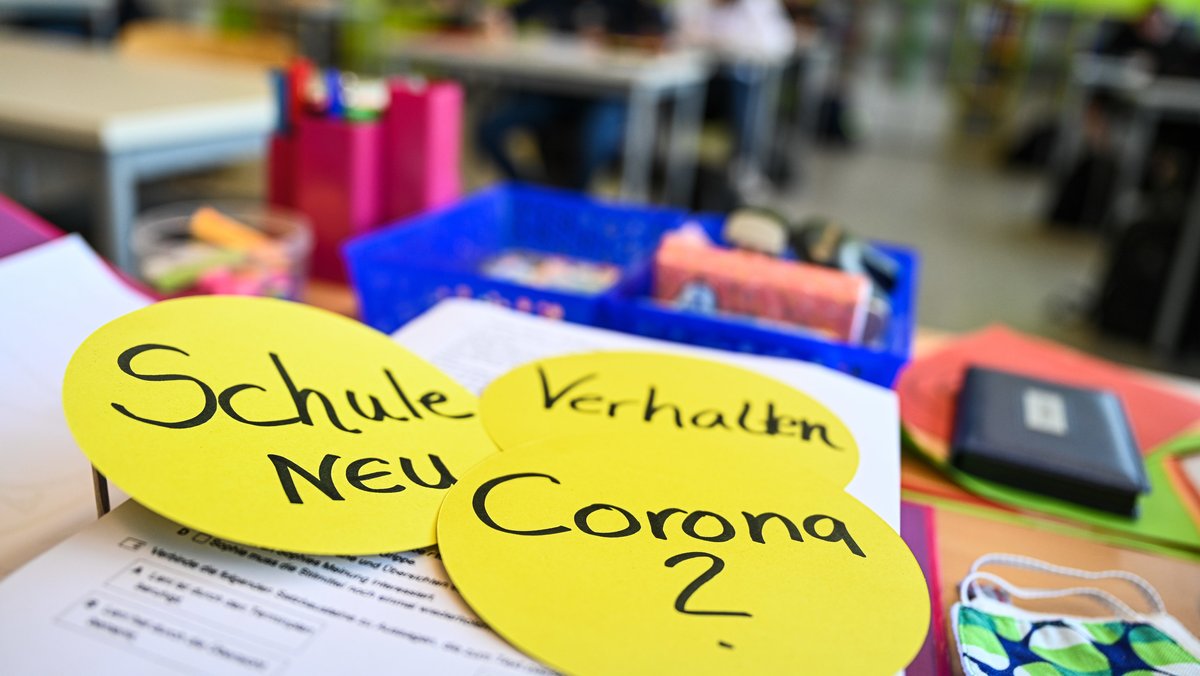 Aufnahme aus einem Klassenzimmer in Baden-Württemberg mit Unterlagen und Papierschildern auf dem Tisch. Aufschrift auf den Schildern: "Corona?", "Schule NEU" und "Verhalten".