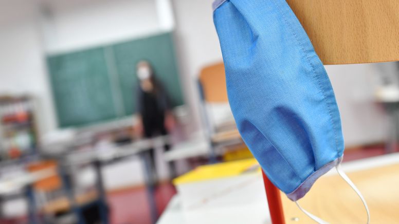 Symbolbild: Eine Maske hängt an einem Stuhl im Klassenzimmer, im Hintergrund steht eine Lehrerin vor der Tafel. | Bild:pa/SvenSimon/Frank Hoermann