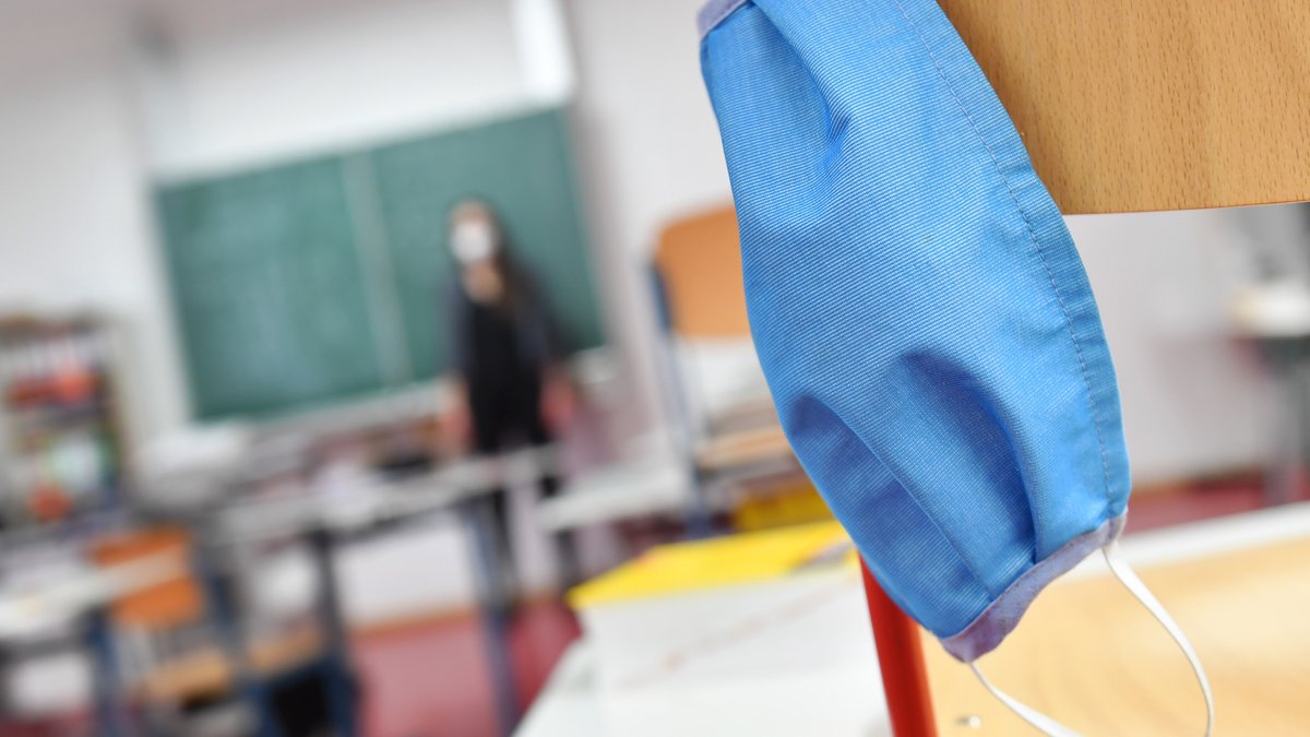 Symbolbild: Eine Maske hängt an einem Stuhl im Klassenzimmer, im Hintergrund steht eine Lehrerin vor der Tafel.