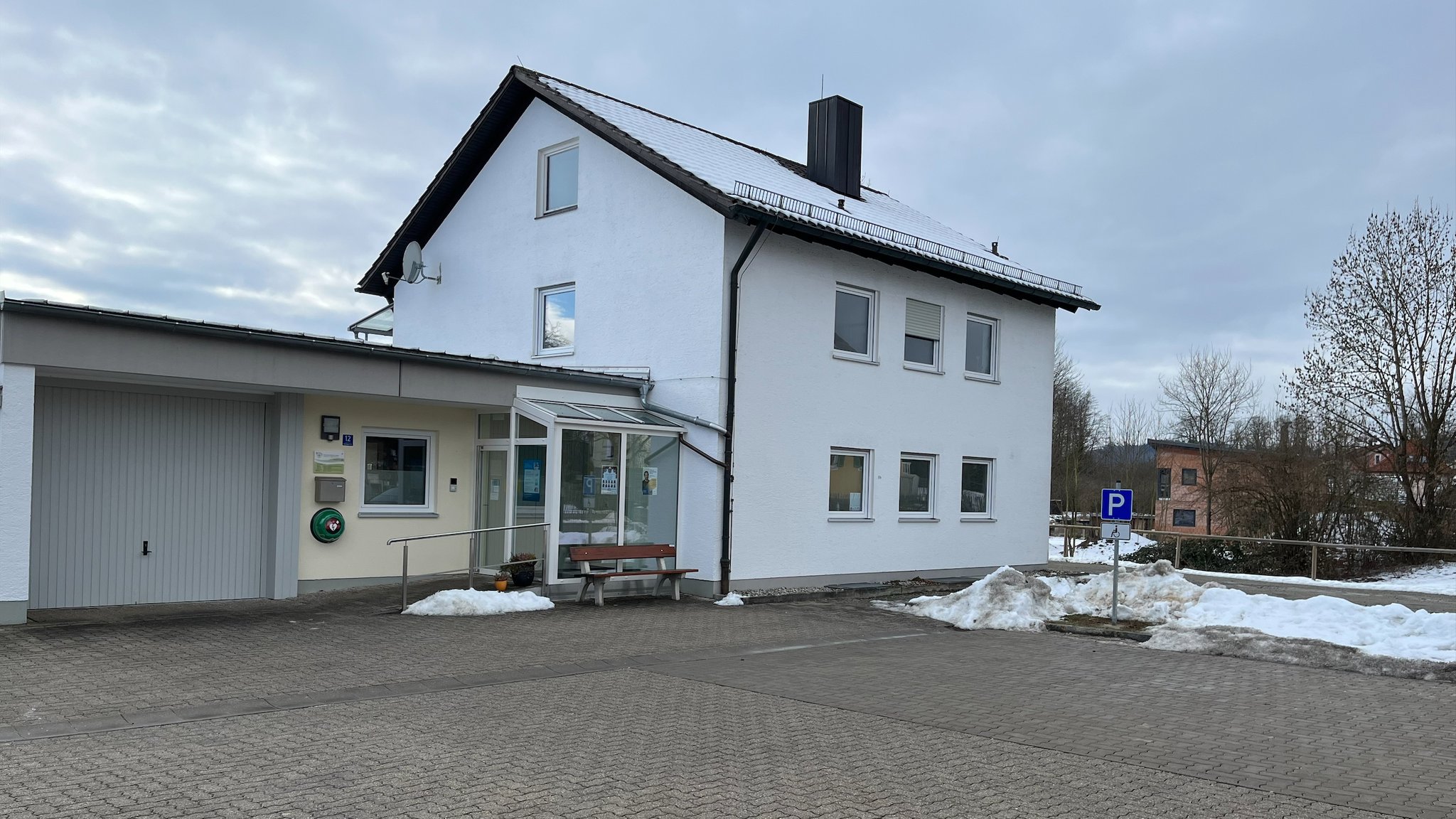 Das Foto zeigt ein freistehendes Haus mit Garage und einem gläsernen Eingangsbereich. Ganz klein ist das Schild mit den Öffnungszeiten der Hausarztpraxis zu sehen. | Bild: BR / Nadine Cibu Das Foto zeigt ein freistehendes Haus mit Garage und einem gläsernen Eingangsbereich. Ganz klein ist das Schild mit den Öffnungszeiten der Hausarztpraxis zu sehen.