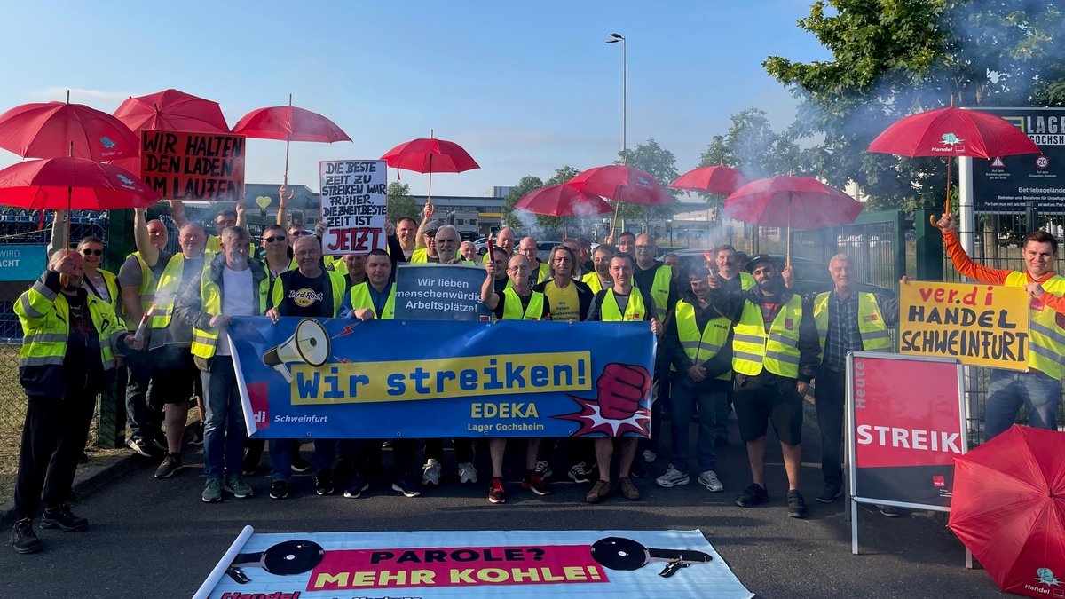 Menschen stehen mit Streikplakaten und roten Schirmen vor dem Edeka-Zentrallager in Gochsheim