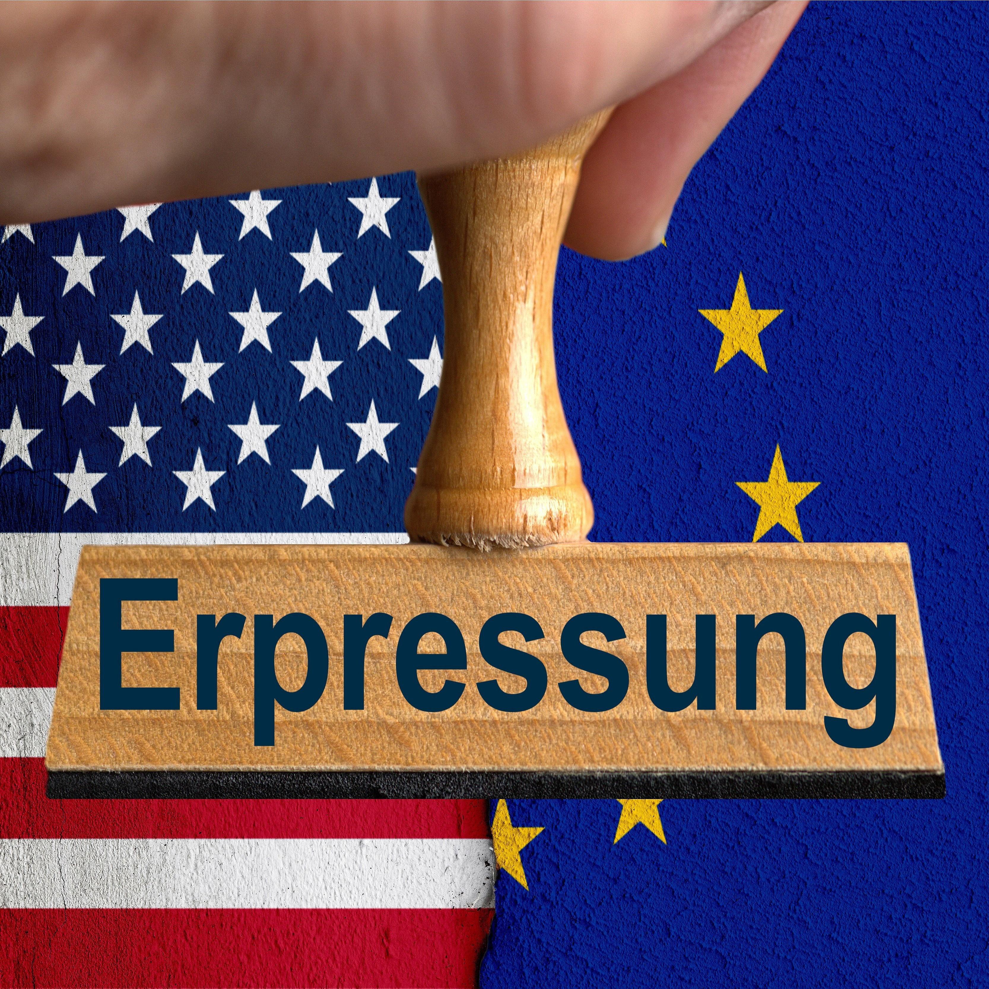 Ein symbolischer Holzstempel mit der Aufschrift "Erpressung", gehalten von einer Hand im Anschnitt vor der US-Flagge und der Flagge der EU auf einer verputzten Wand mit einem vertikalen Absatz im Hintergrund.
