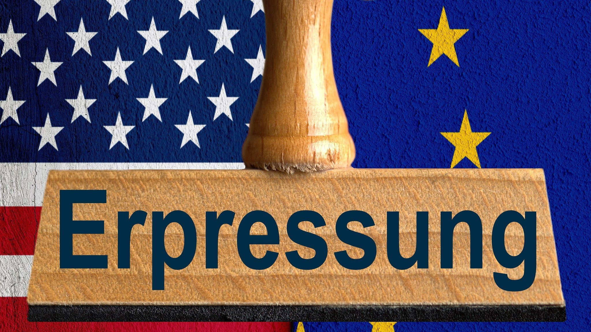 Ein symbolischer Holzstempel mit der Aufschrift "Erpressung", gehalten von einer Hand im Anschnitt vor der US-Flagge und der Flagge der EU auf einer verputzten Wand mit einem vertikalen Absatz im Hintergrund.