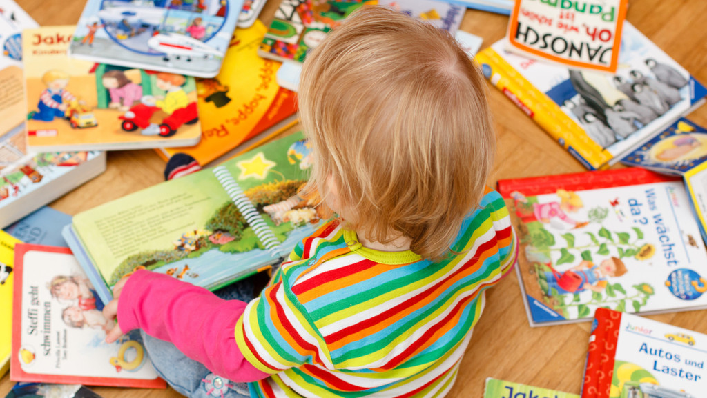 Kleinkind sitzt zwischen Büchern und hat ein aufgeschlagenes Kinderbuch auf dem Schoß