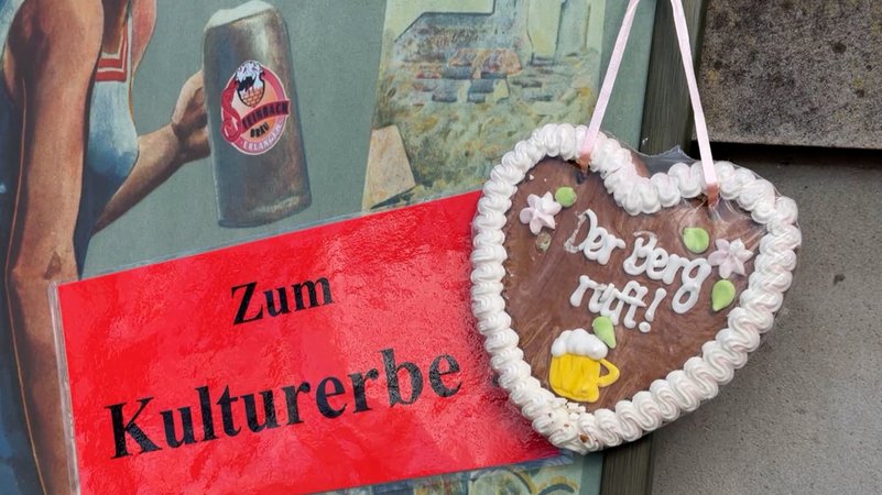 Schild mit Text "Zum Kulturerbe" neben einem Lebkuchenherz mit dem Text "Der Berg ruft". | Bild: BR Schild mit Text "Zum Kulturerbe" neben einem Lebkuchenherz mit dem Text "Der Berg ruft".