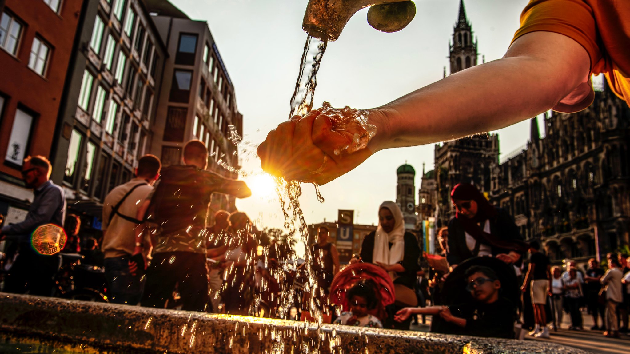 Eine Hand greift in den Wasserstrahl eines Trinkbrunnens in der Münchner Innenstadt, während die Sonne untergeht.