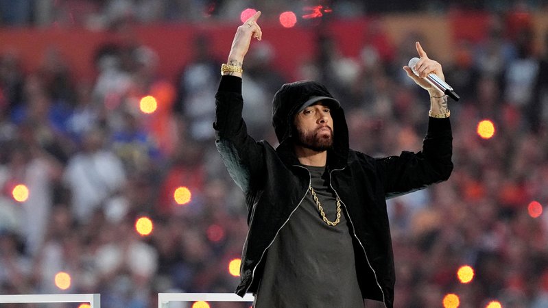 Eminem im schwarzen Hoodie, Kapuze über dem Kopf, mit ernster Mine, die Hände nach oben gereckt, in der einen Hand ein Mikro vor Zehntausenden Zuschauern bei der Halftime Show des Superbowl 2022.