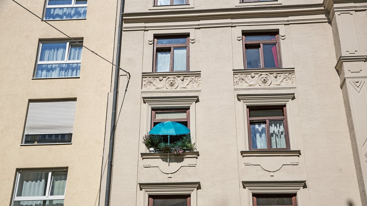 Die Fassade eines Wohnhauses in München. Ein Fenster ist mit vielen Blumentöpfen begrünt, darüber ein blauer Sonnenschirm