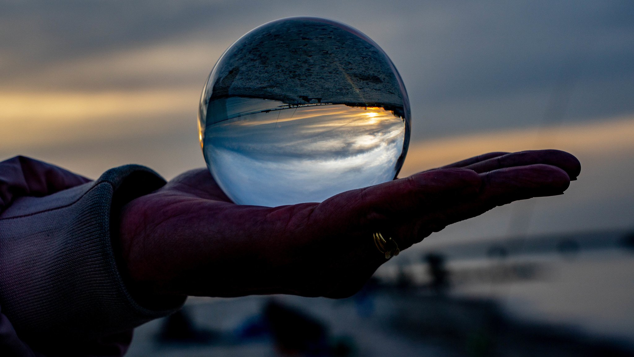 Foto: Eine Hand hält eine Glaskugel auf der Handfläche, darin spiegeln sich der Himmer und die Dämmerung.