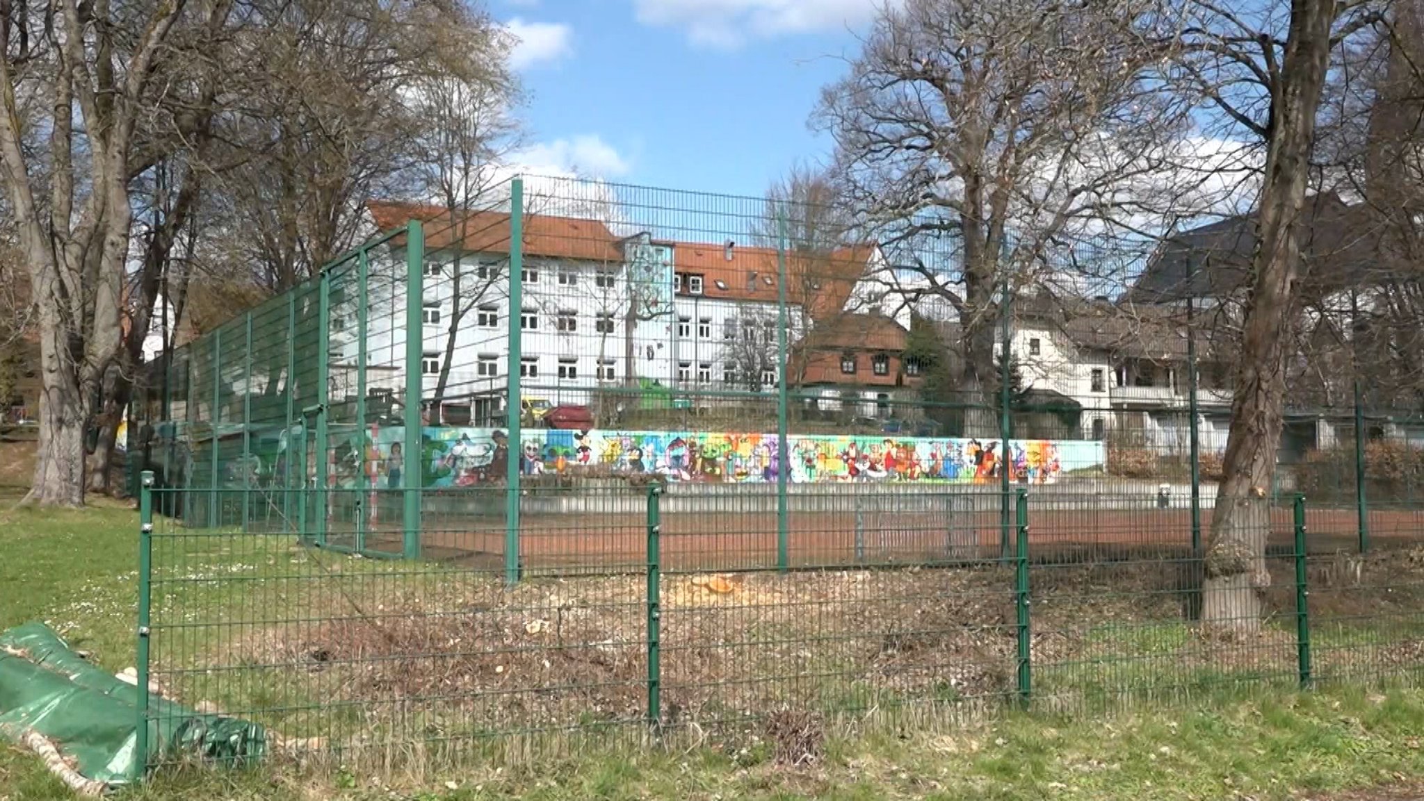 Hinter einem hohen, grünen Zaun befindet sich ein Sportplatz und mehrere große Häuser. 