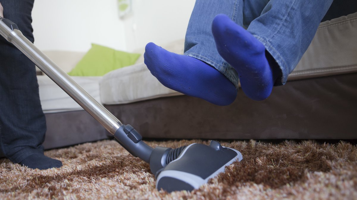 Eine Frau saugt in einer Wohnung einen Teppich mit einem Staubsauger, während eine weitere Person auf dem Sofa sitzt und die Füße hochhebt.