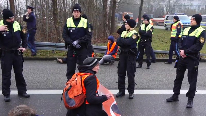 Aktivisten haben sich auf der Ausfahrt der A96 bei München-Sendling auf die Fahrbahn geklebt.  Mehrere Polizisten stehen vor Ihnen.  