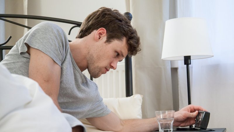 Ein Mann liegt wach in seinem Bett und schaut auf den Wecker auf seinem Nachttisch. Die Hälfte der Erwerbstätigen leidet unter Schlafstörungen.