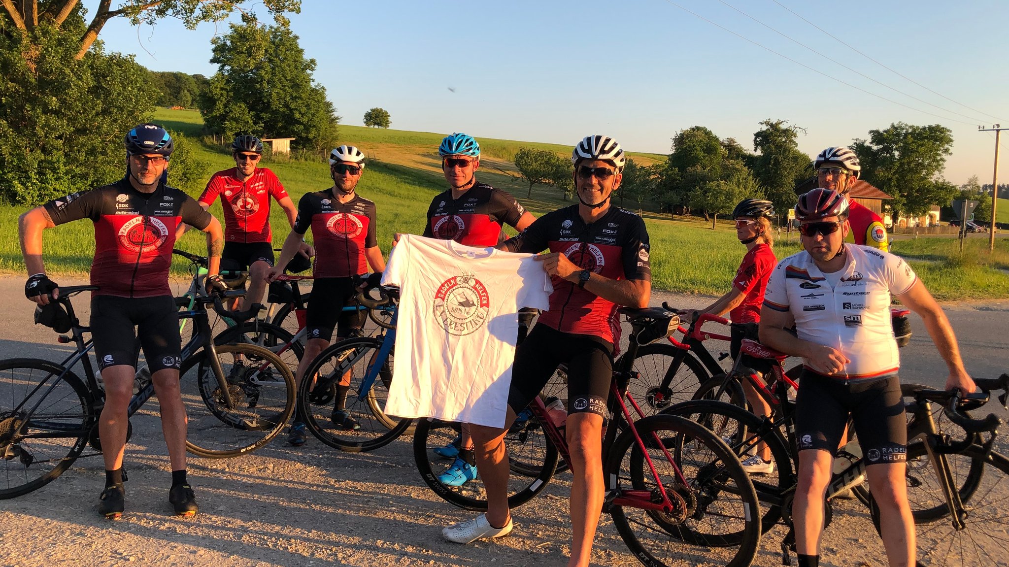 Eine Gruppe von acht Rennradfahrern, der Mann in der Mitte zeigt das Siegertrikot des Vereins "Radeln und Helfen". Das bekommt jeder, der 8.848 Höhenmeter radelt, innerhalb von 24 Stunden.