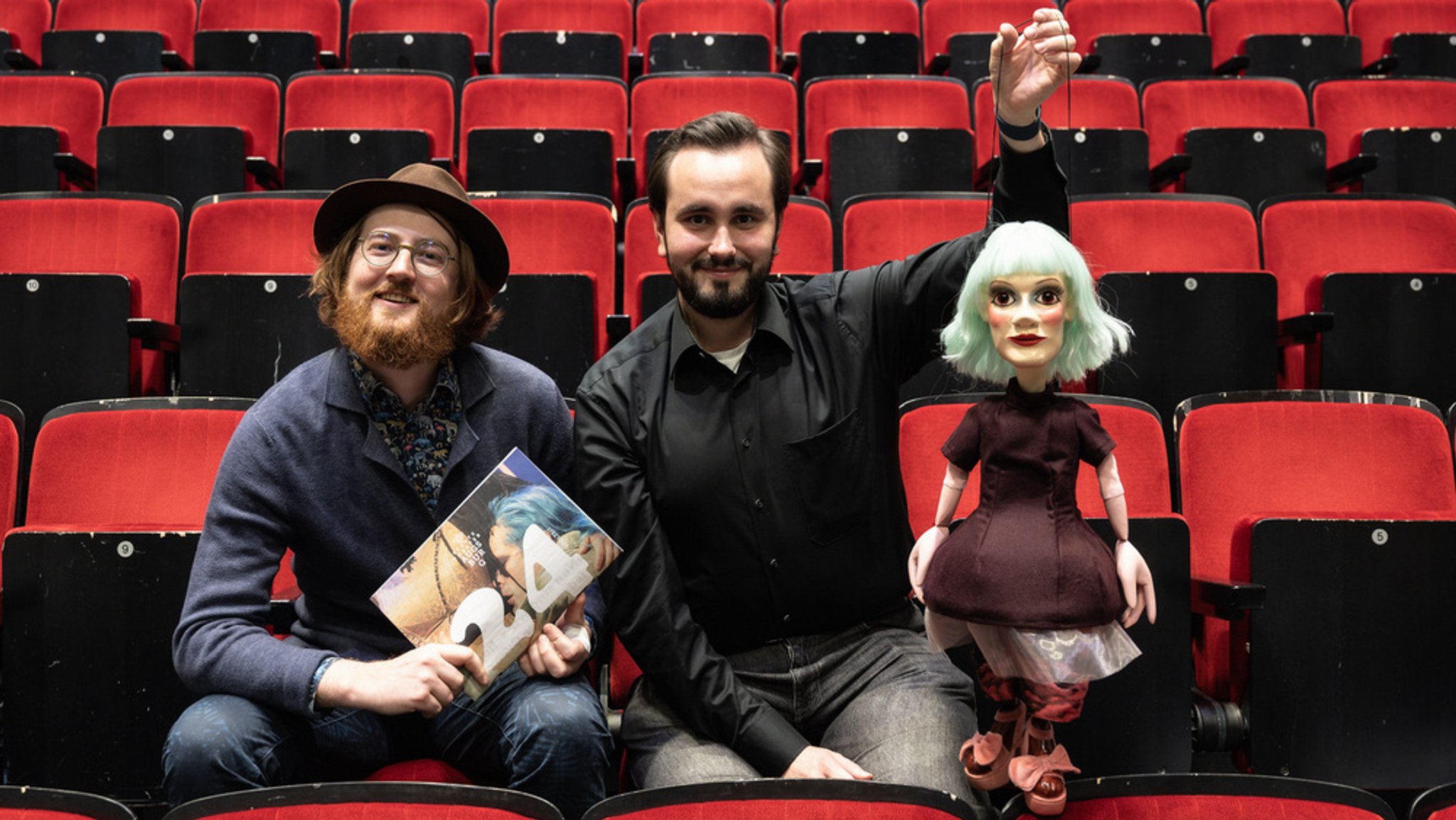 Lukas Joshua Baueregger (Produktionsleitung) und Benjamin Seuffert (Technische Realisation) mit einer Puppenkisten-Marionette und dem Programmheft zur Spielzeit 2024/25 des Staatstheaters Augsburg