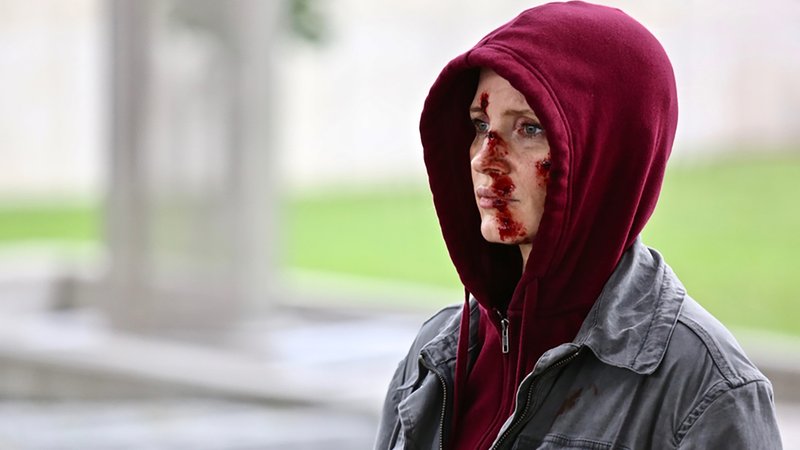 Eine Frau mit blutigen Schürfwunden im Gesicht und einer Kapuze auf dem Kopf steht im Freien (Jessica Chastain in "Code Ava").