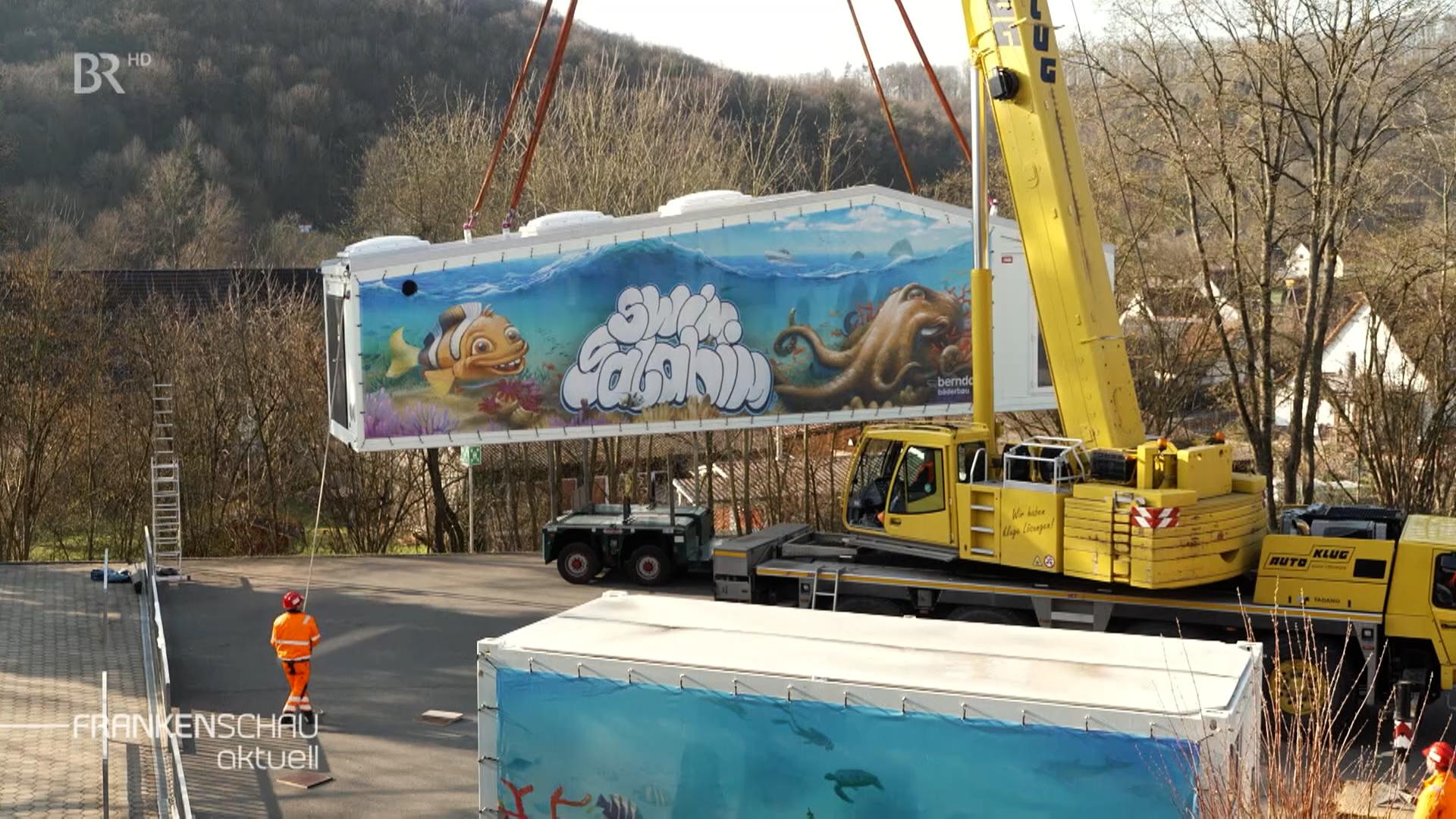 Ein Schwimmbad-Container wird mithilfe eines Krahnes aufgestellt.