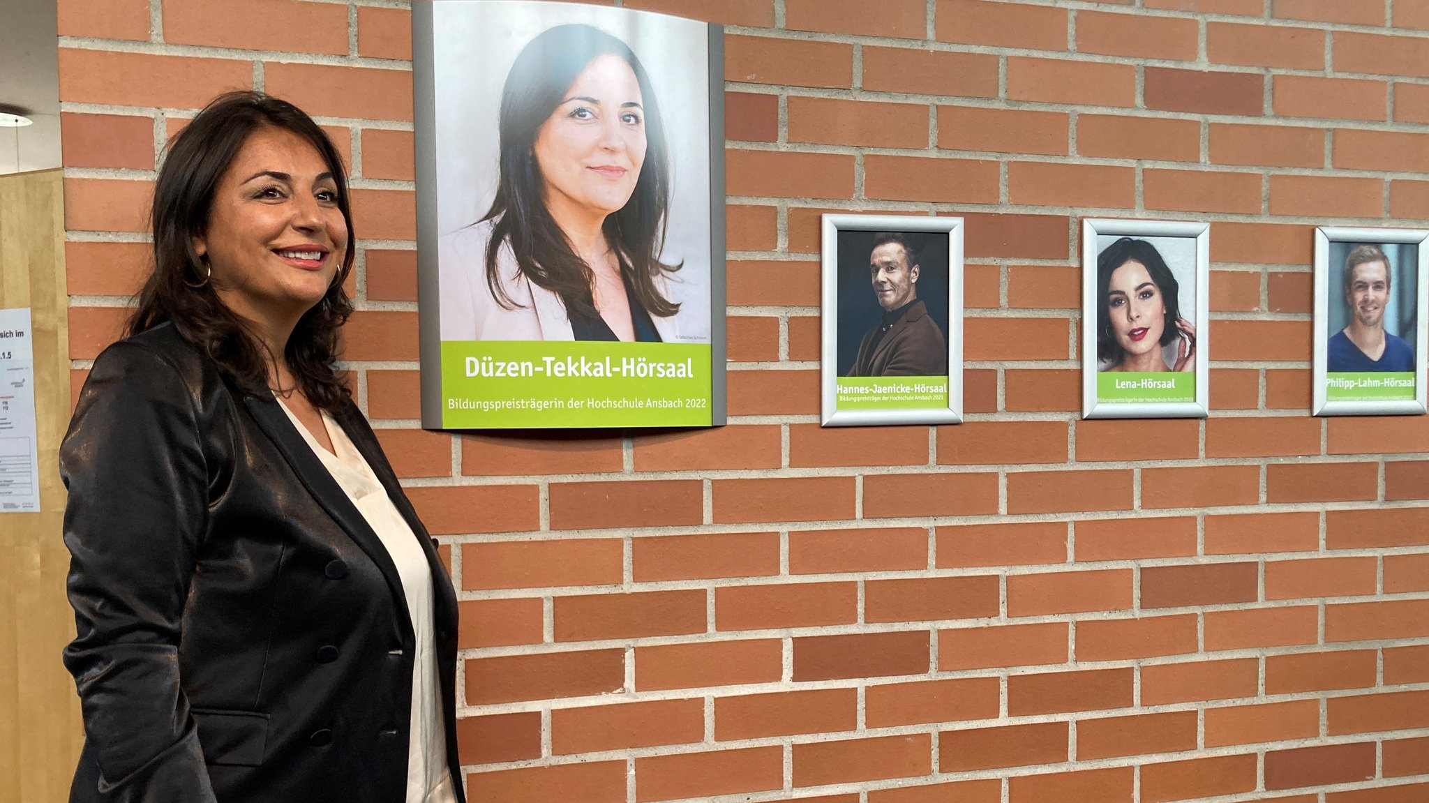 Düzen Tekkal vor einer Wand mit roten Backsteinen, auf der ein Schild mit der Aufschrift "Düzen-Tekkal-Hörsaal" hängt