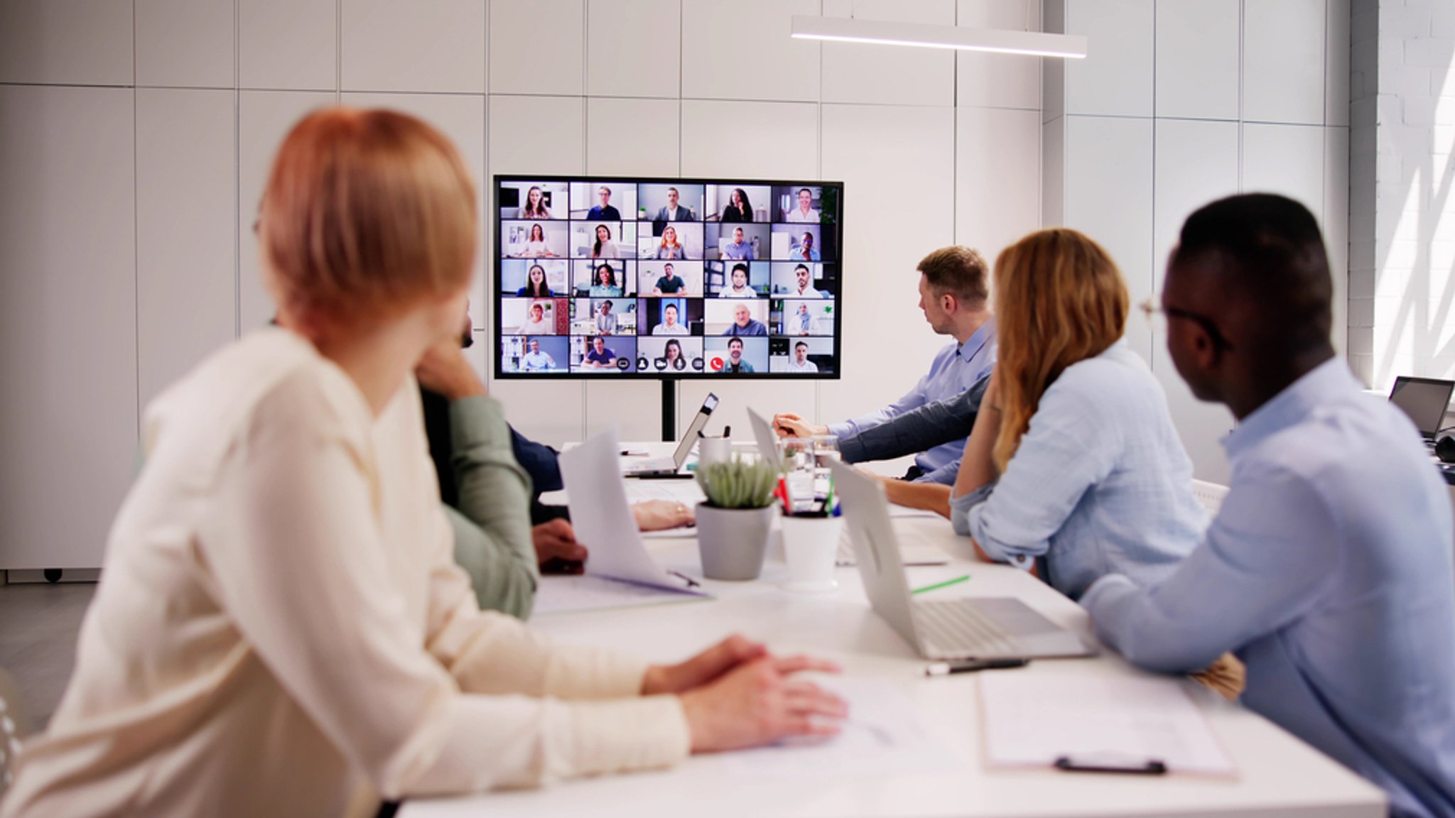 Ein Team sitzt im Konferenzraum und verfolgt eine große Videokonferenz auf einem Bildschirm mit vielen zugeschalteten Teilnehmenden. | Bild: stock.adobe.com/Andrey Popov Ein Team sitzt im Konferenzraum und verfolgt eine große Videokonferenz auf einem Bildschirm mit vielen zugeschalteten Teilnehmenden.