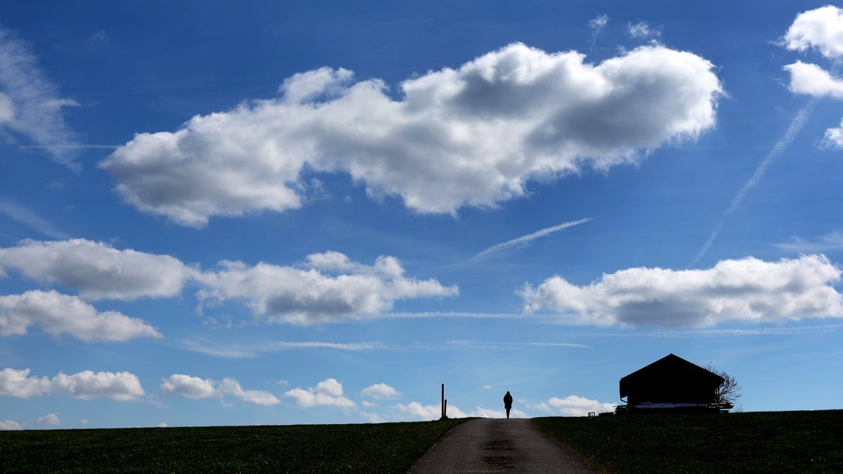 Eine Frau spaziert auf einem Feldweg unter weiß-blauem Himmel.