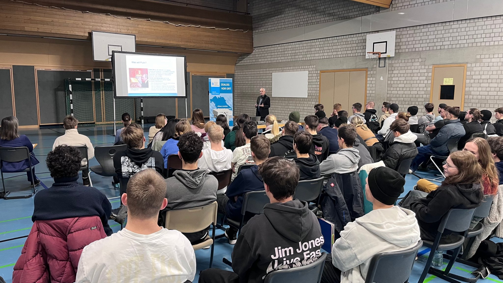 Viele Schüler sitzen auf Stühlen in einer Turnhalle und hören einem Vortrag zu